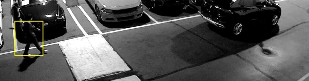 A black-and-white surveillance image captured by an AI-powered security camera using object detection AI to monitor a parking lot. The system highlights a person walking near parked vehicles, demonstrating real-time security monitoring and AI threat detection to reduce false alarms and enhance safety.